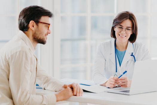 Horizontal shot of doctor and patient discuss something, talk about diagnosis, use laptop computer, pose in consulting room, check medical records. Sick man gets prescription from therapist.