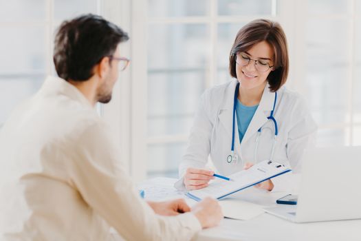 Medical appointment. Female doctor gives professional medical help to male patient, explains written information on paper in clipboard, gives support and good service, pose at hospital near desktop.