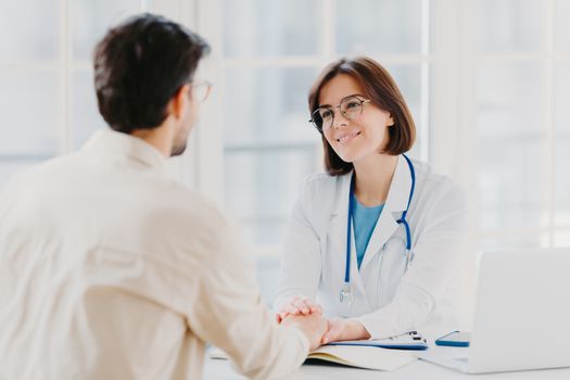 Confident female doctor holds hands of ill patient, persuades everything will be alright, dressed in white medical gown, gives advice, pose in hospial office. Consultation and diagnosis concept