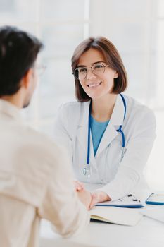 Medical trust concept. Vertical image of female doctor reassures her male patient, hold hand and talk calmly about illness cure, gives professional consultation and support, pose at doctors office