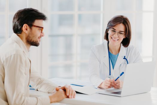 Caring woman doctor gives prescriptions to patient, sit together at table and look at laptop computer, fill up application form online. Sick man visit healthcare clinic, gets medical consultancy