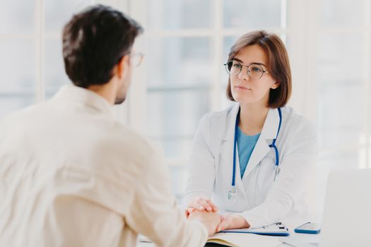 Friendly female doctor tries to support patient, holds his hands, gives useful consultation and explains medical information, makes diagnostic examining, pose in hospital room. Heathcare, assistance