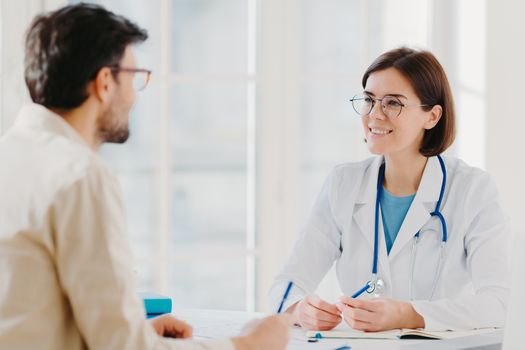 Photo of male patient writes all details makes notes during consultancy with doctor sit in spacious office. Physician tells treatment issues and explains diagnosis to hospital visitor. Medical concept