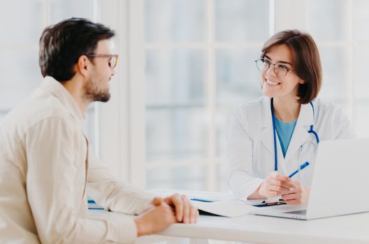 Attentive female doctor explains diagnosis to patient, gives professional consultancy, points with pen at laptop, talk about health concerns, pose at private clinic in modern office, show prescription