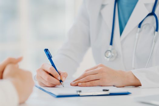 Unrecognizable woman doctor writes down in clipboard, makes prescription for patient, records data for analysis, wears white gown. Close up, focus on hands. Medicine, insurance, health care concept