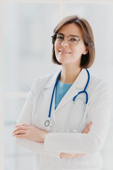 Shot of confident brunette female doctor or surgeon stands with hands crossed, wears white uniform, cares about health of patients ready to give advice any time. People, occupation, profession concept