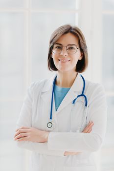 Beautiful cheerful woman doctor wears white gown, glasses and phonendoscope, keeps hands crossed, looks confidently at camera, has short dark hair, stands against white background, works in clinic