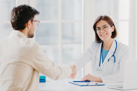 Partnership, assistance, trust and medicine concept. Female doctor shakes hands with thankful patient for good treatment and professionalism, pose in clinic, medical records near on white table