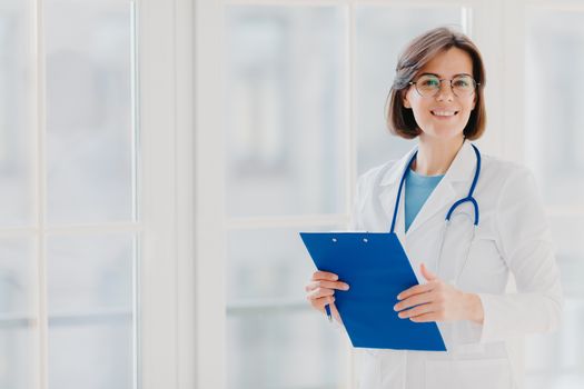 Smiling female doctor stands with clipboard, makes prescription for patinet, wears white gown, poses indoor against big window, works in hospital, holds medical record. Copy space for your information