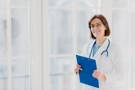 Female cardio therapeutist wears round glasses, white medical coat and phonendoscope, writes with pen in clipboard, stands near big window in own clinic, thinks over patient case and treatment