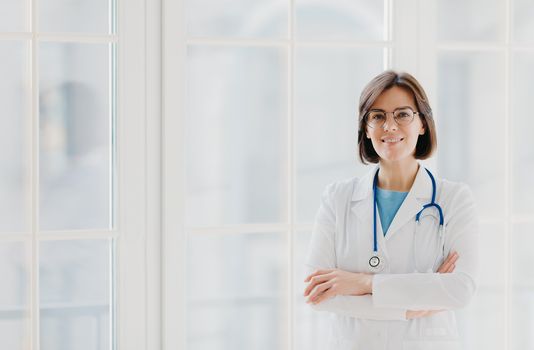 Self confident professional doctor stands with arms crossed, wears white medical gown with stethoscope, thinks about work positively, poses against big window. Healthcare and occupation concept