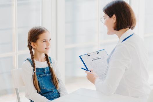 Back view of attentive female pediatrician listens carefully child complaints, writes down notes in personal patient form, gives advice how to cure cold, sit opposite each other in modern clinic