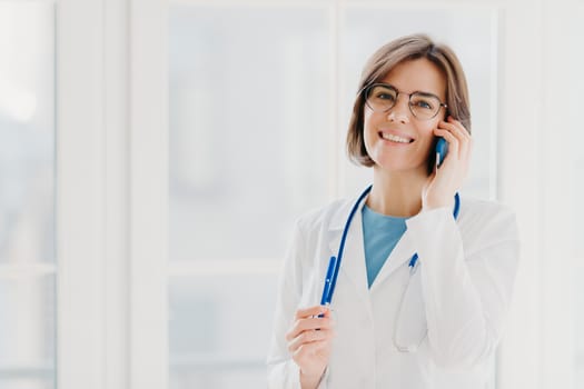 Positive woman doctor consults patient via cellphone, looks gladfully at camera, holds pen, wears white coat, poses in medical office, poses near window. Healthcare and consultation concept.