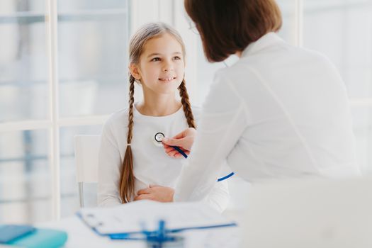 Family general practitioner examines child, listens lungs with stethoscope, makes prescription pose in moren hospital office. Ill kid has cold, comes to pediatrician for good advice has health checkup