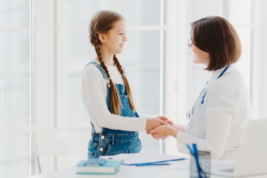 Female doctor shakes hands with girl child patient, talks supportive words, cares about kid during professional consultation, ready always to help with good treatment. Medical insurance concept