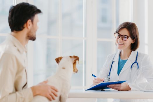 Man brings his pet fot vet examination in clinic, tells symptoms of ill dog. Happy woman veteranian writes down necessary prescription in clipboard for jack russell terrier after careful checkup