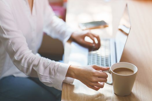 Woman hand on work desk with a laptop computer, a cup of coffee, with morning sunlight.