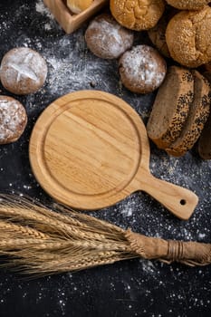 An empty wooden cutting board and wheat on the table with various kinds of bread.