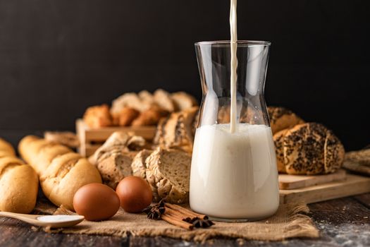 Pour the milk in a glass bottle placed on the sack. A lot of bread and eggs lay on a wooden table.