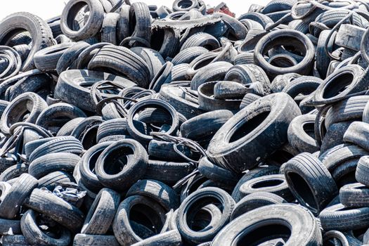 Assorted pile of old and used automotive road tires, showing a variety of tread patterns in a tires shop back yard. No people. Copy space