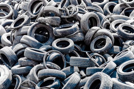 Assorted pile of old and used automotive road tires, showing a variety of tread patterns in a tires shop back yard. No people. Copy space