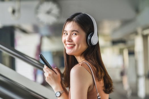 Young Asian woman passenger wearing headphone and using smart mobile phone to listening music and walking up the stairs in subway station when traveling in big city, commuter and transportation concept