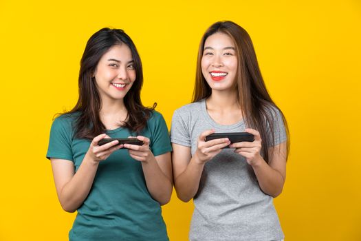 Two Happiness Asian smiling young woman gamer using smart mobile phone and playing games on isolated yellow color background, Lifestyle and leisure with hobby concept