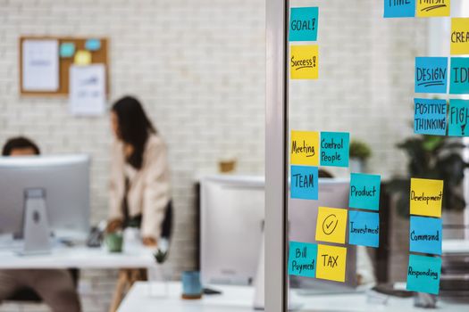 Various post it notes of business strategy wording over the glass wall in office or coworking space, business colleagues working and discussing together, startup team work and small business concept