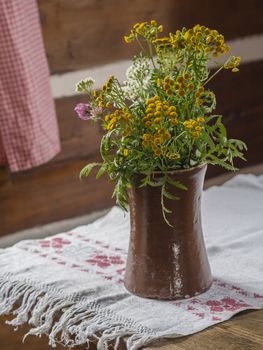old brown ceramic vase with yellow white and pink field flowers on rustic embroider tablecloth on wooden table
