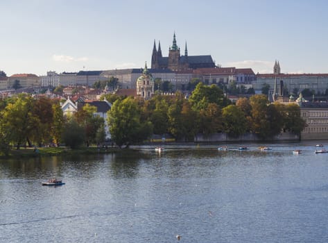 panorama of Prague Castle and St. Vitus Cathedral over Vltava river and Gradchany, Czech Republic, golden hour light, summer sunny day, tourists relaxing on boats.