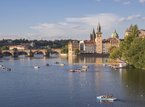 panorama of Charles bridge over Vltava river and houses of Old Town, Prague, Czech Republic, golden hour light, summer sunny day, tourists relaxing on pedal boats.