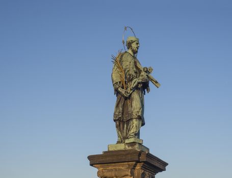 Baroque Bronze Statue of Saint John of Nepomuk with Golden Star Halo holding crucifix with Jesus Christ on Charles Bridge in Prague, Czech Republic, sunny day, clear blue sky.
