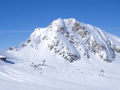 Winter mountains landscape with colorful dressed skiers on white snow slopes. view from the top of Kitzsteinhorn mountain, blue sky. Kaprun ski resort, National Park Hohe Tauern, Austrian Alps, Europe