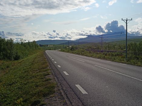 Lonely winding asphalt road and railroad in Abisko village and national park with birch forest and mountains, summer blue sky clouds.