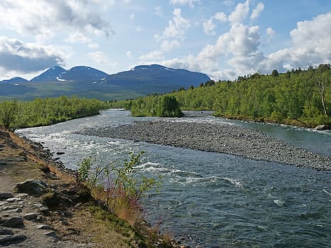 Beautiful wild Lapland nature landscape with blue glacial river, birch tree bushes, snow capped mountains and dramatic clouds. Northern Sweden summer at Kungsleden hiking trail