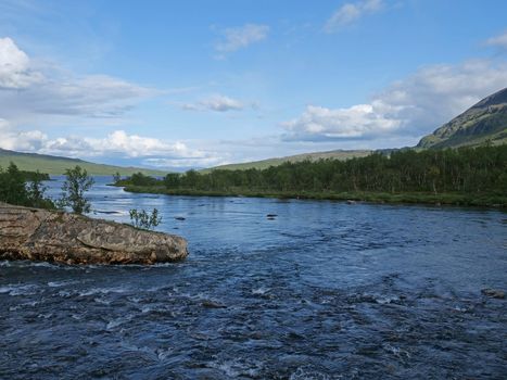 Landscape with a blue glaciar river Abiskojokk, rock, birch tree and green hills in Abisko National Park, Lapland, Northern Sweden, Kungsleden trail. Summer sunny day.
