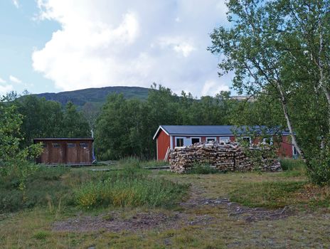 Red building of AbiskoJaure STF mountain hut. Green hills, birch forest on the Kungsleden Trail, summer blue sky.