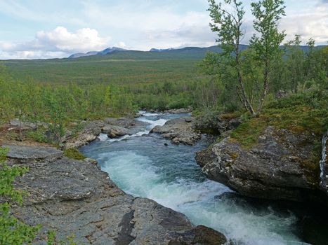 Landscape with rapids of wild blue glaciar river Kamajakka, rock, birch tree, green mountains at Rovvidievva sami village, Lapland, Northern Sweden, Summer sunny day.