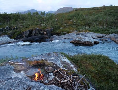 Lapland landscape with burning campfire and rapids of wild river Kamajakka, rock, birch trees and mountains at Rovvidievva sami village, Northern Sweden, Summer evening.