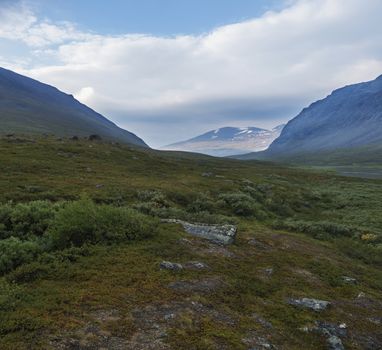 Beautiful northern landscape with wild glacier river Kamajokk, boulders and spruce tree forest in Kvikkjokk in Swedish Lapland. Summer sunny day, golden hour, dramatic clouds.