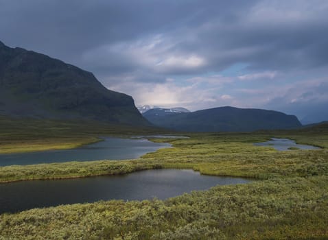 Beautiful wild Lapland nature landscape with blue glacial river, birch tree bushes, snow capped mountains and dramatic clouds. Northern Sweden summer at Kungsleden hiking trail