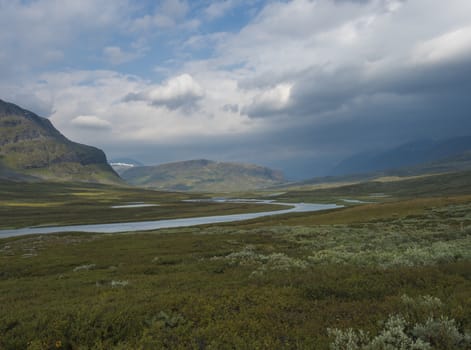 Beautiful wild Lapland nature landscape with blue glacial river, birch tree bushes, snow capped mountains and dramatic clouds. Northern Sweden summer at Kungsleden hiking trail