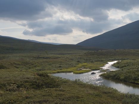 Beautiful wild Lapland nature landscape with blue glacial river, birch tree bushes, snow capped mountains and dramatic clouds. Northern Sweden summer at Kungsleden hiking trail