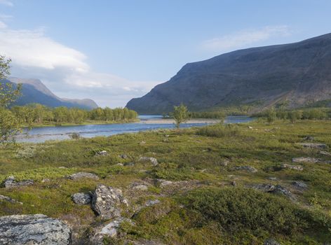 Beautiful wild Lapland nature landscape with blue glacial river, Kaitumjaure lake, birch tree forest and mountains. Northern Sweden summer at Kungsleden hiking trail. Blue sky background.