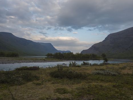 Beautiful wild Lapland nature landscape with blue glacial river, Kaitumjaure lake, birch tree forest and mountains. Northern Sweden summer at Kungsleden hiking trail. Blue sky dramatic sunset clouds
