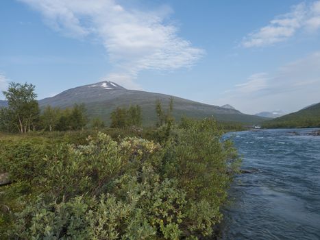 Beautiful wild Lapland nature landscape with blue Tjaktjajakka river, Kaitumjaure, birch tree forest and mountain Sanjartjakka. Northern Sweden summer at Kungsleden hiking trail. Blue sky background.