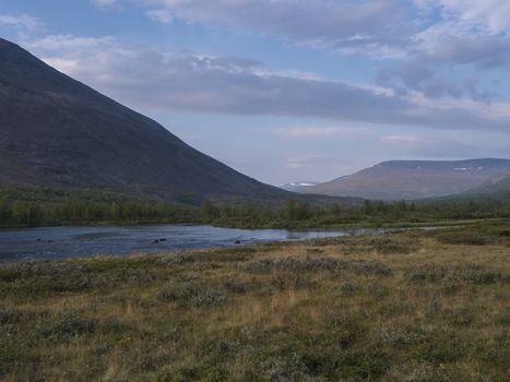 Beautiful wild Lapland nature landscape with blue Tjaktjajakka river, Kaitumjaure, birch tree forest and mountain Sanjartjakka. Northern Sweden summer at Kungsleden hiking trail. Blue sky background.