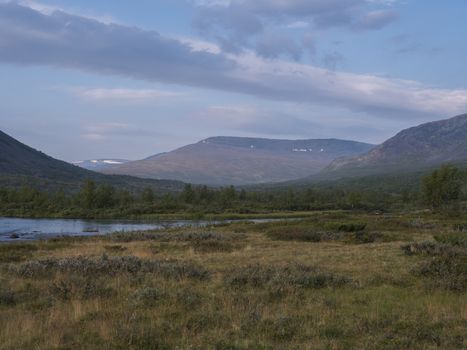 Beautiful wild Lapland nature landscape with blue Tjaktjajakka river, Kaitumjaure, birch tree forest and mountain Sanjartjakka. Northern Sweden summer at Kungsleden hiking trail. Blue sky background.