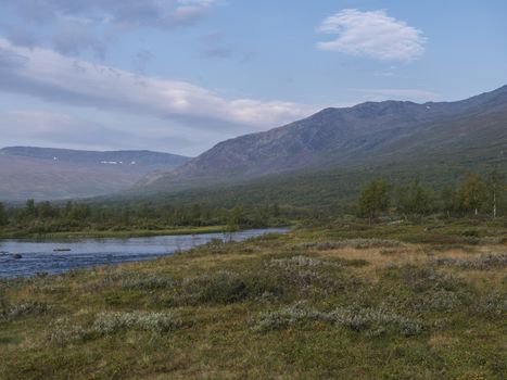 Beautiful wild Lapland nature landscape with blue Tjaktjajakka river, Kaitumjaure, birch tree forest and mountain Sanjartjakka. Northern Sweden summer at Kungsleden hiking trail. Blue sky background.