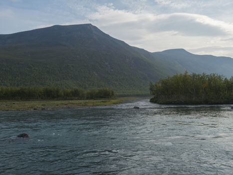 Confluence of wild Tjaktjajakka river and Kaitumjaure lake with small birch tree forest island and mountains. Lapland nature landscape in summer at Kungsleden hiking trail
